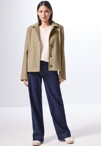 Lyhyt oliivinvihreä takki, jossa on läpät ja viisi nappia, yhdistettynä vaaleaan villapaitaan ja leveälahkeisiin tummansinisiin farkkuihin, joita on käytetty beigen väristen tennareiden kanssa.