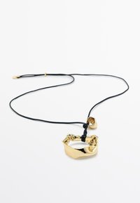 Ciondolo geometrico in oro su collana con cordoncino nero. Il ciondolo ha una superficie strutturata con una forma irregolare, che ne aumenta l'interesse visivo.