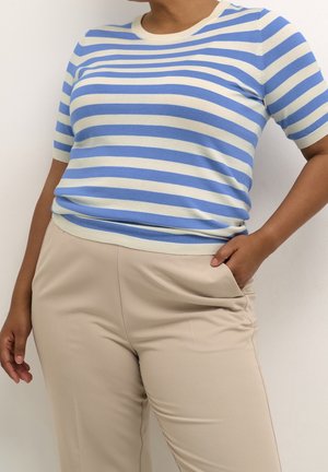 Vrouw draagt blauw-wit horizontaal gestreept kort mouwen topje en beige hoge taille broek, met één hand in de zak, tegen een witte achtergrond.