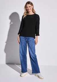 Schwarzer Strickpullover mit strukturiertem Muster, kombiniert mit weiten blauen Jeans und weißen Sneakers, vor einem einfarbigen hellen Hintergrund.