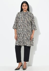Tunika con stampa leopardata, maniche a tre quarti e colletto, abbinata a pantaloni neri e décolleté a punta con dettaglio a fiocco.