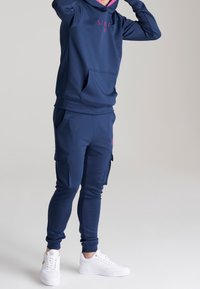 Marinblå huvtröja med rosa logga, framficka, i kombination med matchande joggerbyxor med sidofickor och ribbade manschetter; vita sneakers.