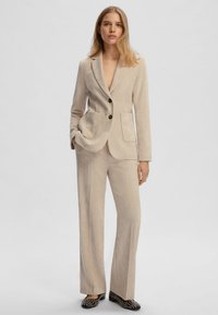 Blazer in corduroy beige con revers a scendere, tasche frontali e due bottoni. Abbinato a pantaloni a gamba larga coordinati e scarpe nere con motivo.