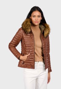 Bruine gewatteerde jas met een capuchon met bontafwerking, ritszakken en geribde manchetten, gecombineerd met een tan coltrui en witte broek.