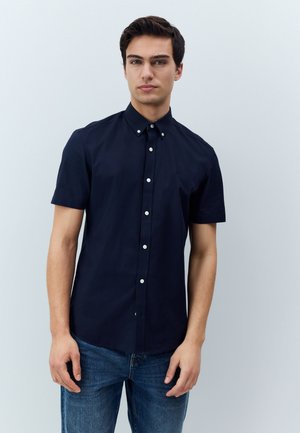 Junger Mann, der vor einem einfarbigen Hintergrund steht, trägt ein geknöpftes navyblaues Kurzarmhemd und blaue Jeans, blickt nach vorne.