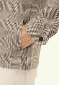 Brown and white checkered jacket with a textured fabric, featuring a side pocket and a black button on the cuff. أبرزDetails visible.