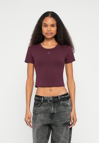 T-shirt cropped cor borgonha com decote redondo e mangas curtas. Apresenta o logótipo "HUGO" à frente. Combinada com jeans cinzentos com efeito desgastado e cinto preto.