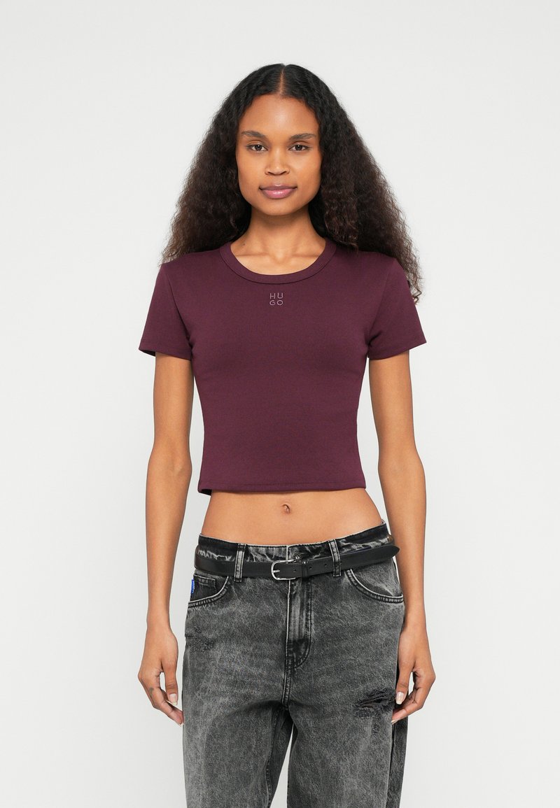 T-shirt cropped cor borgonha com decote redondo e mangas curtas. Apresenta o logótipo "HUGO" à frente. Combinada com jeans cinzentos com efeito desgastado e cinto preto.