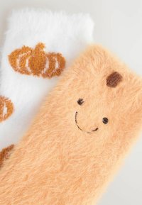 Zwei flauschige Socken mit herbstlichen Motiven: Eine pfirsichfarbene Socken mit einem lächelnden Gesicht und die andere weiße Socken mit einem orangefarbenen Kürbismuster.
