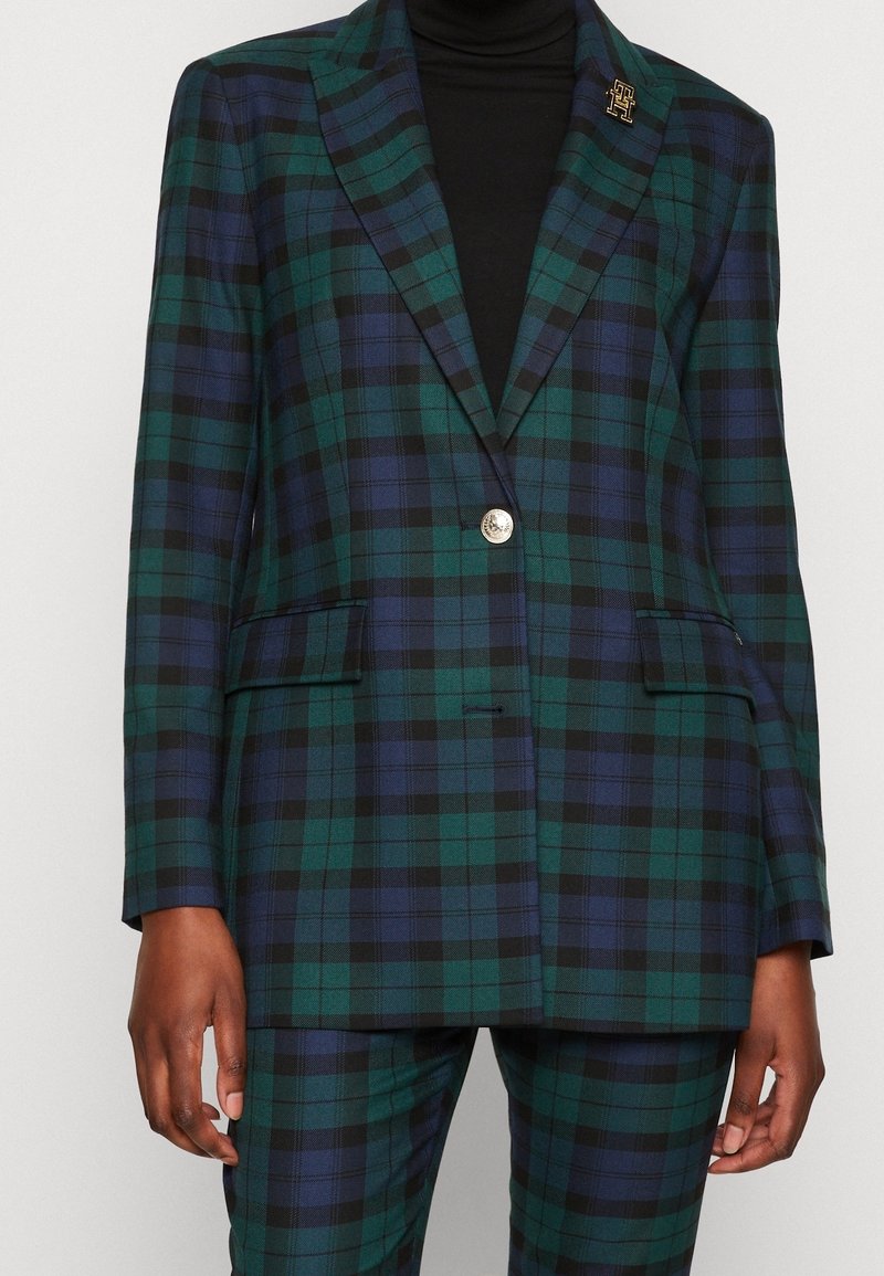 Femme portant un blazer à carreaux verts et bleu marine assorti à un pantalon, avec un col roulé noir en dessous.