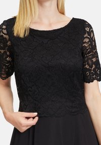 Zwarte kanten top met korte mouwen, bloemenpatroon, geschulpte randen en een ronde halslijn, gedragen boven een effen zwarte rok.