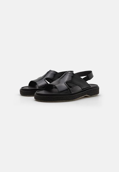 Sandales synthétiques noires avec design découpé, bout ouvert et sangle de talon réglable. Semelle texturée pour une adhérence et un confort optimal.