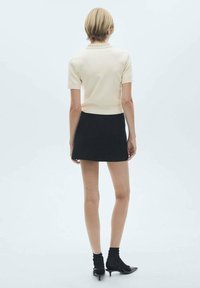 Eine Frau steht mit dem Rücken zur Kamera und trägt ein cremefarbenes Kurzarm-Oberteil, einen schwarzen Minirock, schwarze Socken und schwarze, flache Schuhe auf einem weißen Hintergrund.