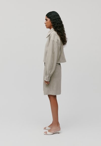 Vrouw met lang krullend haar, gekleed in een beige cropped jack, een bijpassende rok, witte open teen hakken en gouden ringoorbellen, staand in profiel.