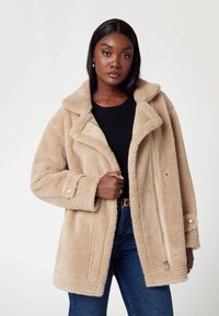 Bež jakna iz umetnega krzna z širokim ovratnikom, sprednjim zadrgo in gumbi na manšeta, teksturirana površina in velikodušna prileganje. Nosena čez črno majico.