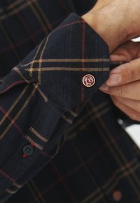 Cămașă din flanel navy cu un model în carouri în nuanțe de aur și roșu. Prezintă un buton rotund de culoare aurie cu un design embossat pe mansetă.