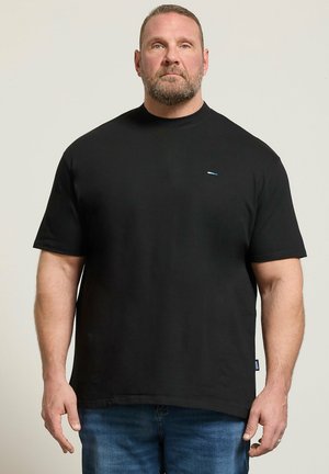 Mann mit kurzen Haaren und Bart steht frontal, trägt ein schlichtes schwarzes Kurzarm-T-Shirt und blaue Jeans vor beigem Hintergrund.