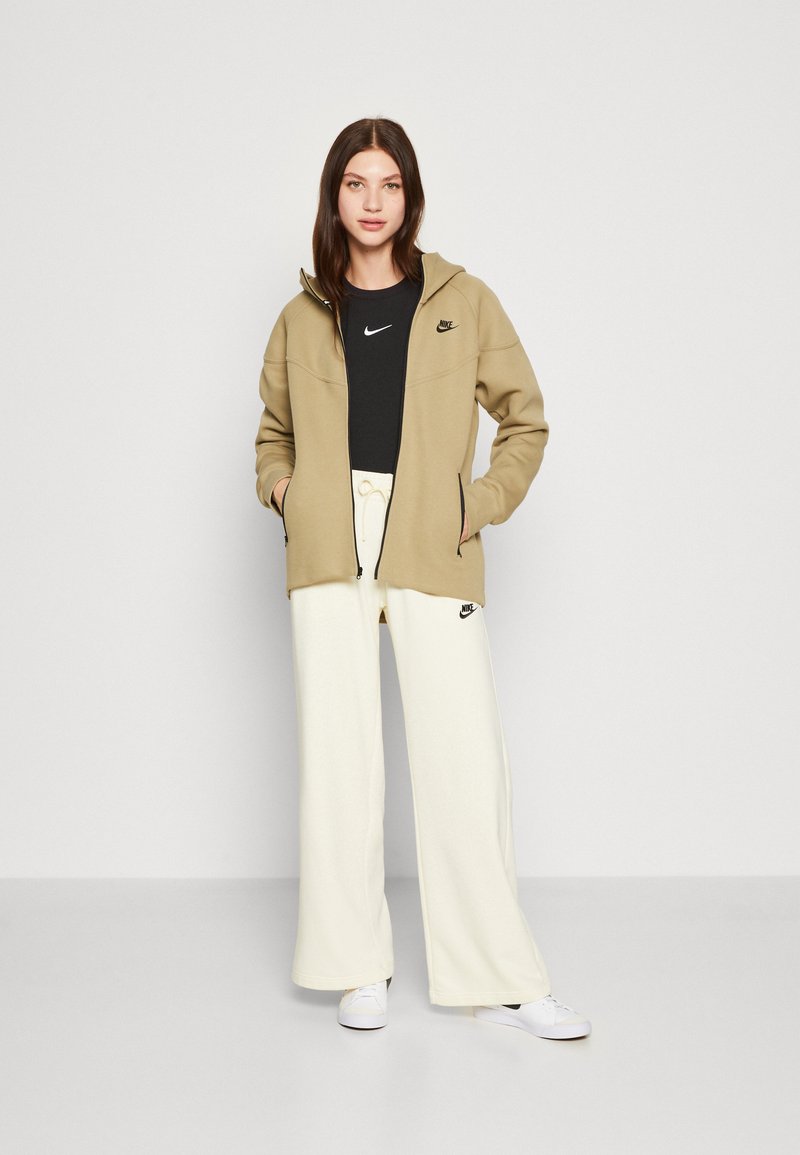 Khakifarbene Zip-Hoodie über einem schwarzen T-Shirt, kombiniert mit weit geschnittenen, cremefarbenen Sweatpants. Mit Nike-Logos auf dem Hoodie und den Pants.