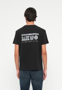 Muž v černém tričku Tommy Jeans se textem "Bank of NYC 1985" na zádech, v kombinaci s tmavě modrými džíny, stojící proti bílému pozadí.