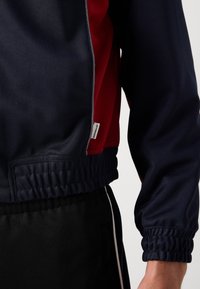 Ναυτικό και κόκκινο αθλητικό μπουφάν με ελαστικές μανσέτες και στρίφωμα, λείο ύφασμα και μια μικρή λευκή ετικέτα στην πλευρική ραφή.