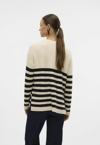 Maglione a righe crema e nero con vestibilità ampia, caratterizzato da un orlo asimmetrico e una texture a costine. Indossato con jeans scuri.