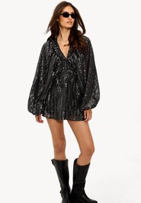 Robe noire pailletée avec un décolleté en V, des détails froncés et des manches bouffantes, associée à des bottes noires jusqu'aux genoux. Lunettes de soleil portées en accessoire.