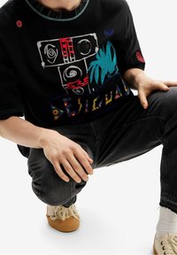 Persona con una camiseta gráfica negra con diseños abstractos y jeans oscuros, agachada con zapatillas beige sobre un fondo blanco.