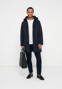 Marineblaue Kapuzenparka mit Reißverschluss, zwei Seitentaschen, kombiniert mit einem weißen Hemd und schwarzen Schuhen, hält eine schwarze Tasche.