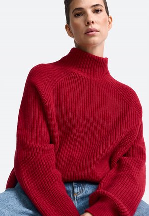 Roter gestrickter Pullover mit hohem Kragen, gerippter Textur und überschnittenen Schultern. Kombiniert mit hellblauem Denim für einen Kontrast.