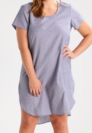 Femme portant une robe courte violette claire à manches courtes et longueur genou avec un petit motif à pois, une main sur la hanche et l'autre détendue le long du corps.