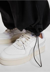 Hvide læder sneakers med grå accenter, der har runde snørebånd, en struktureret sål og en forlænget tunge, parret med sorte baggy bukser.