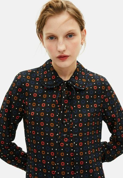 Czarna bluzka w kwiatowy wzór w odcieniach czerwieni, niebieskiego i białego. Posiada okrągły kołnierz, zapięcie na guziki z przodu oraz detal w postaci wiązania przy szyi. Długie rękawy.