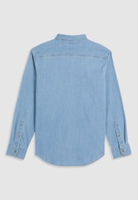 Chemise en denim bleu clair à manches longues posée à plat montrant le dos, avec poignets boutonnés et une couture en empiècement sur le haut du dos.