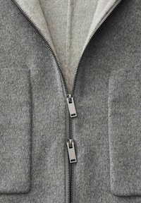 Veste en laine grise avec deux curseurs de fermeture éclair en métal à l'avant et grandes poches rectangulaires de chaque côté.