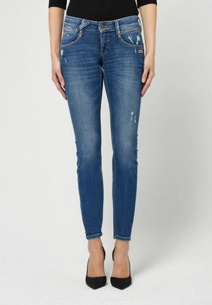 Vrouw draagt slim fit blauwe spijkerbroek met lichte slijtage en zwarte hoge hakken, staand tegen een effen achtergrond.
