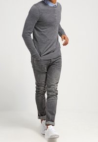 Grau pullover mit runder Ausschnitt, über einem blauen Hemd mit Kragen getragen. Kombiniert mit hellgrauen Jeans und weißen Sneakers. Schlichte Gestaltung, lässige Passform.