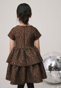 Barna leopárdmintás ruha rövid ujjakkal, rétegzett szoknyával és hátul gombos záródással. Csillogó díszítések és táncdömping a háttérben.