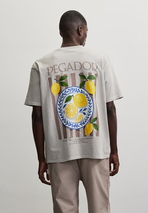 Mand vendt væk iført en beige t-shirt med teksten "PEGADOR" og grafik af citroner og en blå mønstret tallerken på ryggen, kombineret med lyse bukser.