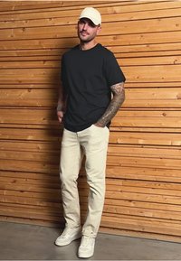 Černé tričko, světle béžové kalhoty a bílé tenisky. Osoba stojí proti dřevěnému pozadí, má na hlavě světlou čepici.