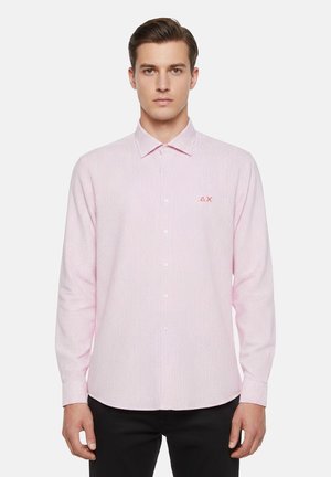 Uomo che indossa una camicia attillata a righe rosa e bianche con bottoni e maniche lunghe, e pantaloni neri, in piedi davanti a uno sfondo bianco.