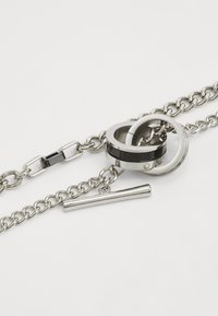 Bracelet en chaîne argentée avec des anneaux entrelacés, une bande accent noire et argentée, et un détail en barre cylindrique lisse.