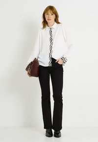 Blusa blanca con ribete negro en forma de concha y puños estampados, combinada con pantalones negros de campana y un bolso burdeos. La modelo está de pie frente a un fondo blanco.