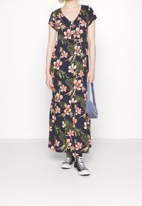 Robe longue fleurie à manches courtes et col en V. Fond sombre avec des fleurs roses et des feuilles vertes. Stylée avec des baskets noires et un sac bleu.
