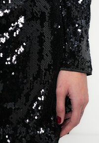 Melna samta audums ar spīdīgiem apaļiem fliteriem, kas pilnībā pārklāj virsmu. Roku ar tumši sarkanu manikīru atbalsta pret audumu.