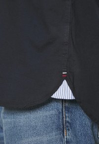 Ciemnoniebieska koszula z zaokrąglonym dołem, z widoczną u dołu paskowaną podszewką. Materiał wydaje się gładki, z delikatnymi detalami przeszycia.