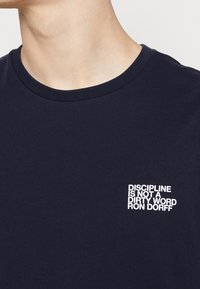 Mörkblå bomullströja med rund halsringning. Har vit text: "DISIPLIN ÄR INTE ETT SMUTSIGT ORD RON DORFF" på vänster bröst.