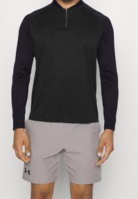 Man som bär en svart och mörkgrå långärmad träningströja med en kort dragkedja samt ljusgrå shorts från Under Armour.