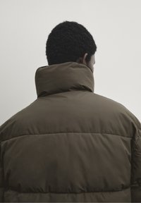 Bruine gewatteerde jas met een hoge kraag, met een doorstikte textuur en een matte afwerking. De achterkant benadrukt een brede, gestructureerde silhoutte.