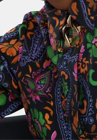 Multikleurige overhemd met knopen op een zwarte basis, met bloem- en paisleypatronen in oranje, roze, groen en paars, en een gouden accent.