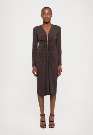 Robe longue marron à manches longues avec un décolleté en V et une fermeture éclair à l'avant, présentant des détails froncés le long du corps. Associée à des sandales noires à lanières.
